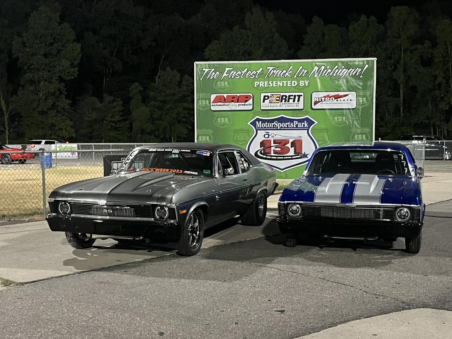 Two classic cars parked on a track with a large green sign in the background.