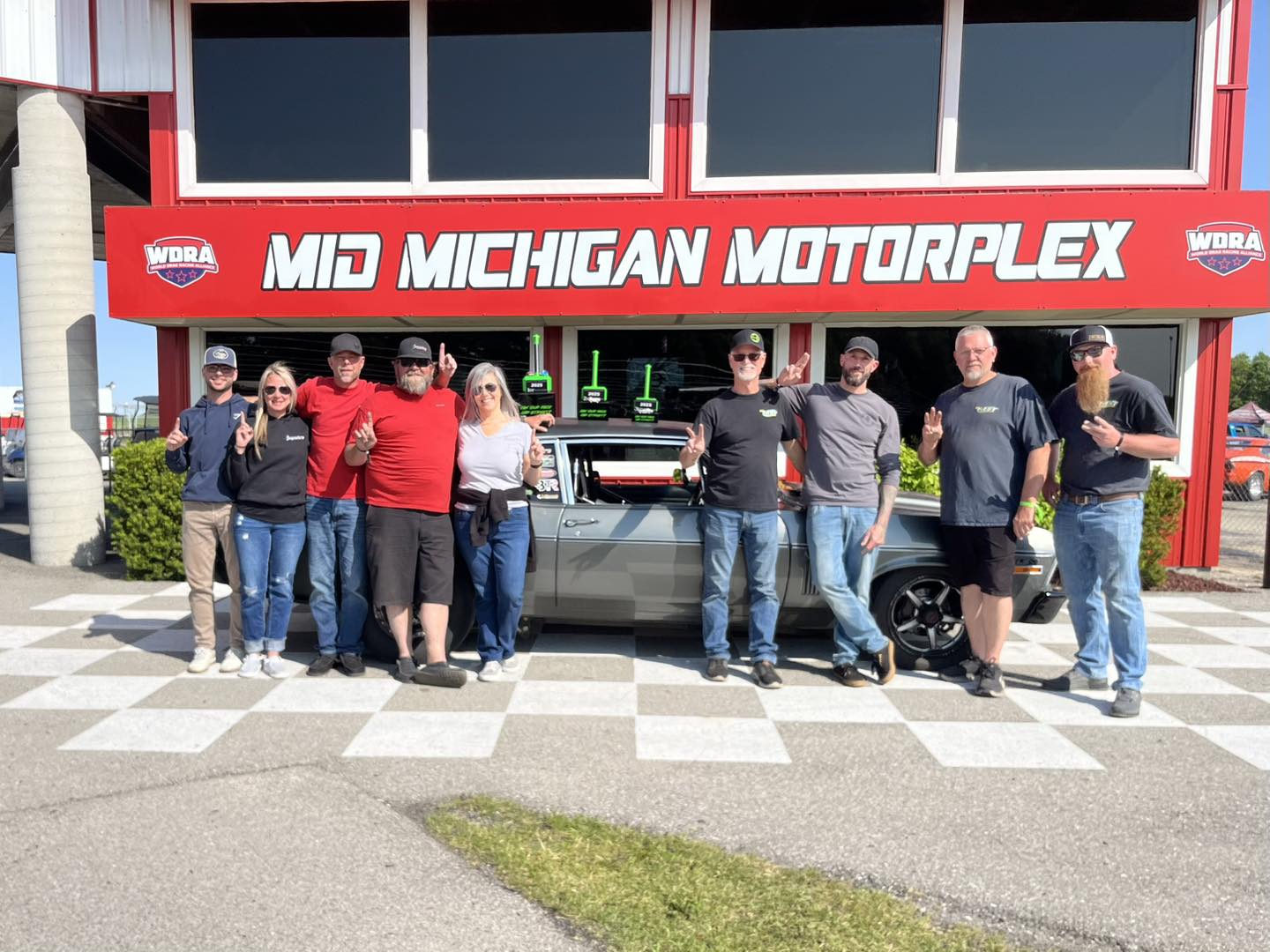Group of people posing in front of the Mid Michigan Motorplex building.
