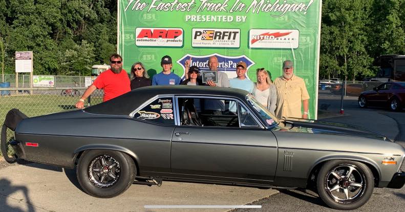 Group of people posing with a classic car in front of a promotional backdrop.